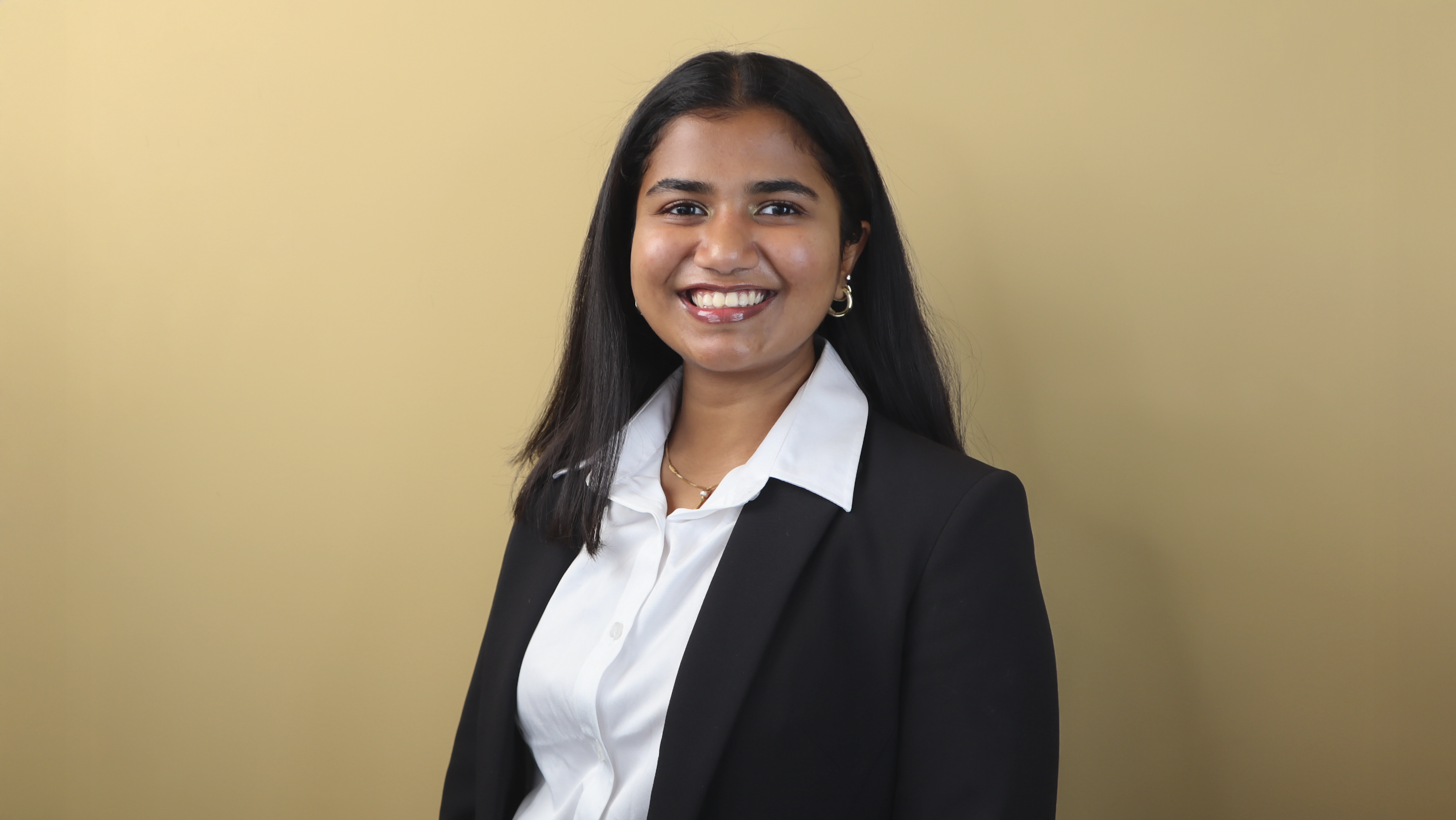 Janani Kamalakkannan wears a black blazer over a white button up while smiling in front of a beige background. Janani Kamalakkannan wears a black blazer over a white button up while smiling in front of a beige background.